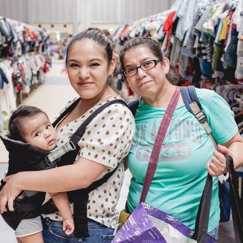 Three generations shop together at their local JBF sale. Mom carries baby in a baby carrier while grandmom stands behind them.